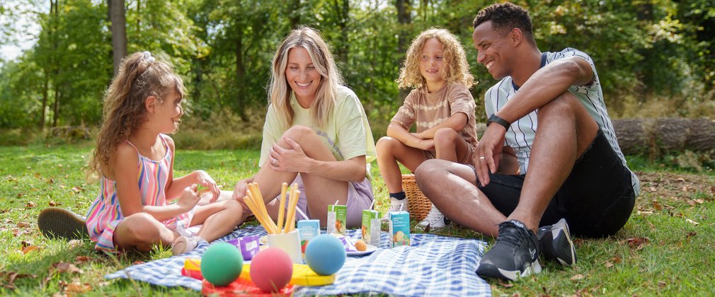 A family of two adults and two children in the forest, sitting with a picnic on a picnic rug. They are smiling at each other. 