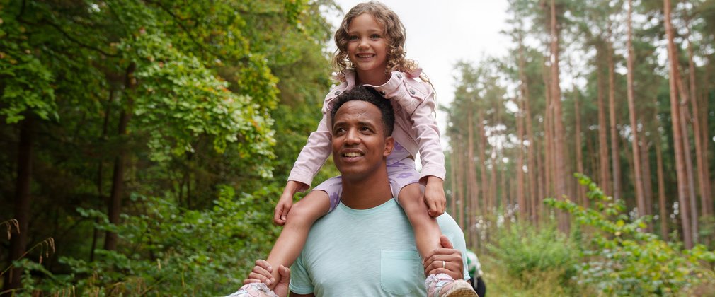 A young, smiling girl being carried on the shoulders of a man, walking along a forest path