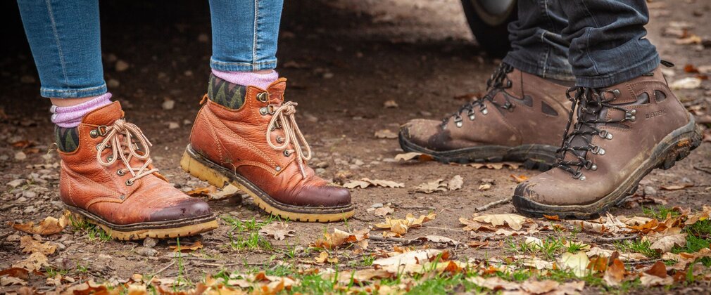 Close-up of two people's walking boots stood on forest ground.