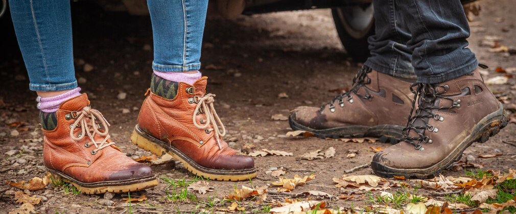 Close-up of two people's walking boots stood on forest ground.