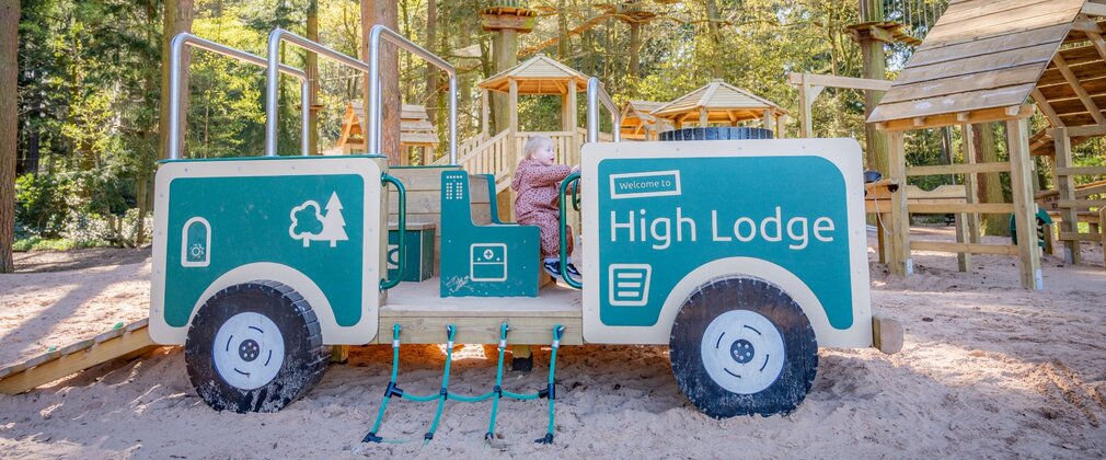 Young girl sat in a wooden truck play structure in a sandy play area.