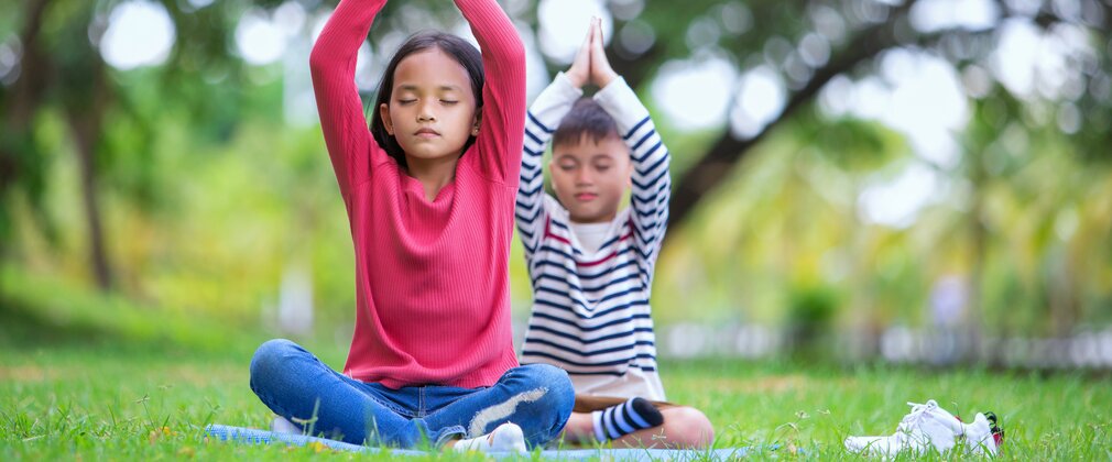 2 children sit crossed legged in a woodland environment with eyes closed and hands over their heads