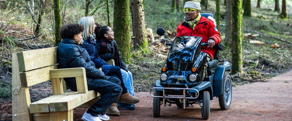 A man on an all-terrain mobility scooter talks to a woman and two children sitting on a bench in the forest.