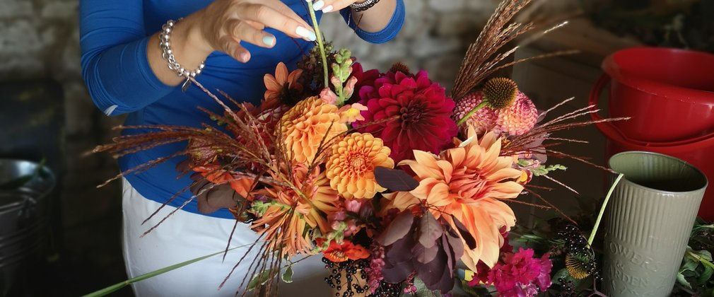 A woman in a bright blue top is arranging vibrant orange and red flowers into a vase