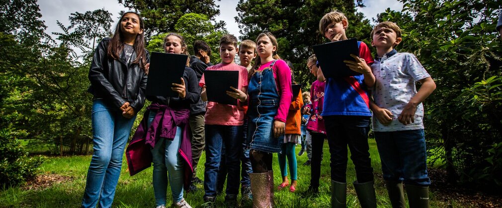 A group of school children, holding clipboards, standing in green space.