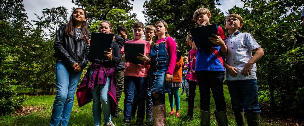 A group of school children, holding clipboards, standing in green space.