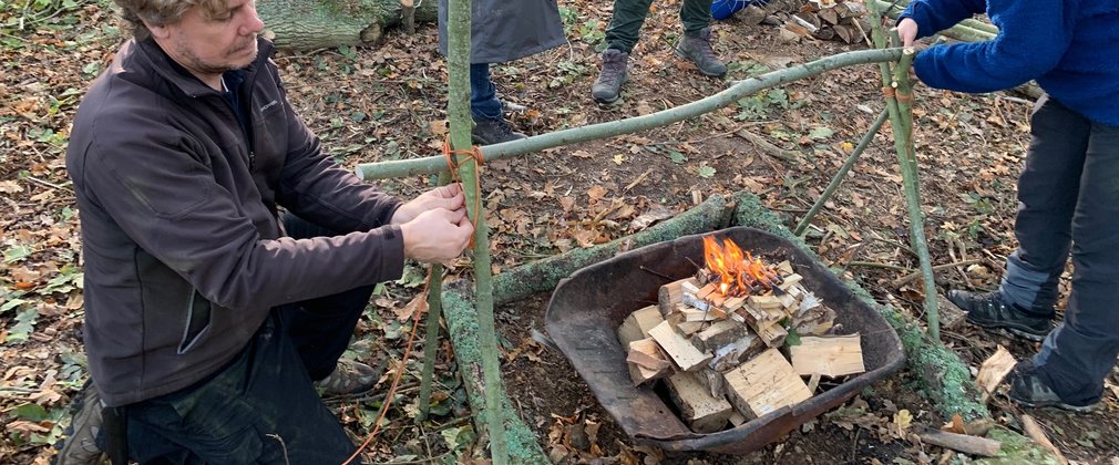 Two people making a wooden tripod over a fire for cooking