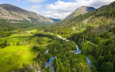 A drone shot of a wooded valley on a sunny day, with a river running through it and mountains behind