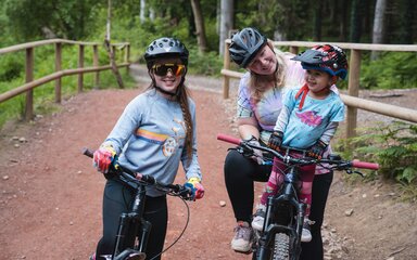 A woman and two young girls on mountain bikes smiling at the camera, ready to head out on a cycling trail.