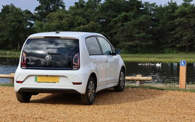 A white car parked in an accessible bay in a gravel car park beside a pond in the New Forest