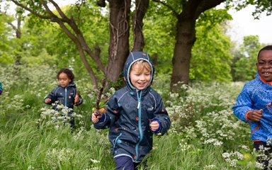 Three children in field with forest backdrop. One child is holding a stick.