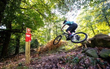 Mountain bike rider doing a jump with a red waymarker sign to his left