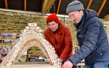 2 gentlemen dressed in winter coats and hats look intently over a wooden arched structure.