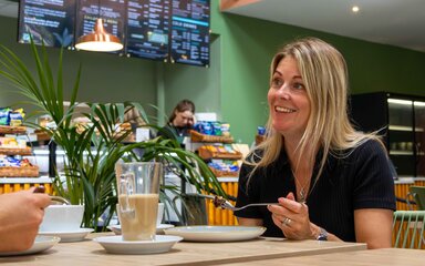 A person sitting inside a cafe smiling at a table with coffee and cake. The cafe counter and menu can be seen in the background.