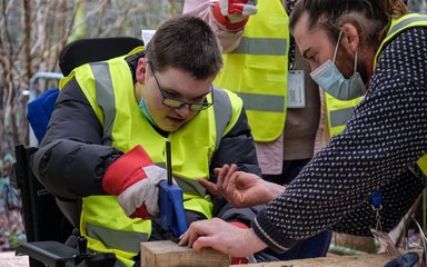 a young male in a wheelchair in a high vis jacket and gloves looks down whilst learning to operate a clamp. A male adult looks on feeding instructions on how to do it.