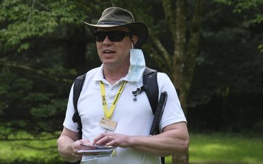 A gentleman in a white short sleeved top, dark glasses and a trilby hat speaks to an audience. He holds a book which contains braille to prompt his sensory walk.