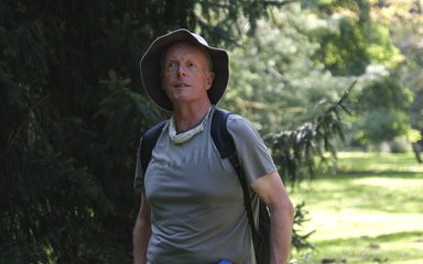 A gentle man wearing a grey t-shirt and a sun hat speaks to an audience on a sensory guided walk