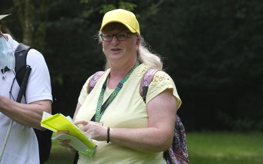 A woman dressed in an yellow top with a yellow baseball hat and glasses speaks to an audience in front of her. 