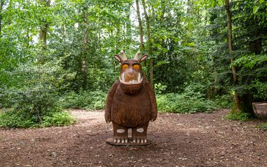 A large wooden sculpture of the children's book character the Gruffalo. Large and Brown with horns, orange eyes and two large fang teeth.