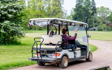 A small shuttle bus drives away from the camera with two visitors waving in the back.
