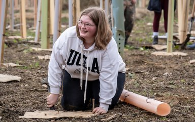 A young lady smiles as she hammers in a peg to a brown piece of matting where she will be planting a baby tree.