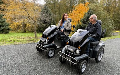 A man and a woman driving two all terrain mobility scooters through a forest