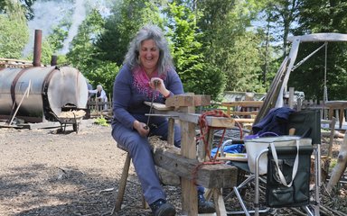 A woman dressed in a long sleeve purple top with a pink scarf sits astride a shave horse - a large wooden structure. She holds a draw knife - a long shaving tool - which she cuts into the wooden block.