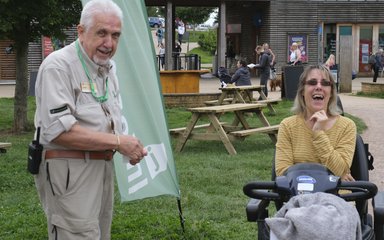 A gentleman volunteer dressed in a light khaki shirt and trousers smiles while distributing a Mobility scooter to a woman in a yellow stripped top, who is laughing.