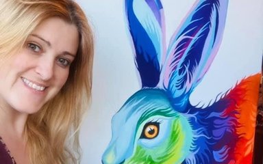 A woman with long blonde hair holds a large painting of a painting she has done of a rabbit made up of rainbow colours. She smiles as she looks at the camera.