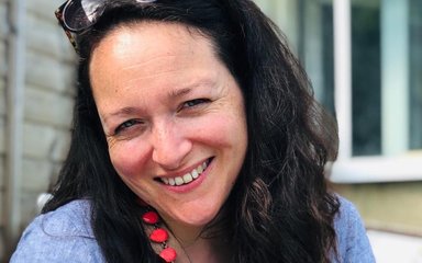 A woman with long dark hair smiles at the camera. She wears sunglasses on her head and has an orangy pink necklace on over a light blue cotton dress