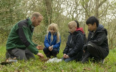 A group of children kneels on the grass with a uniformed ranger, looking at something is his hand.