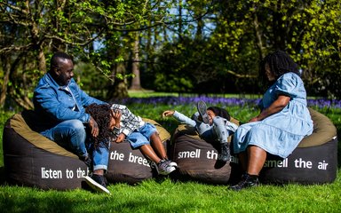 A family sit on large bean bags that have words saying listen to the birds, smell the air, feel the breeze. the children laugh and fall back on the beanbags, while the parents look relaxed and smile. 