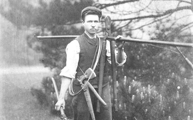 Black and white archive photo of a man wearing a flat cap and carrying gardening tools.