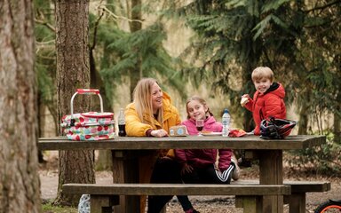 Mother and two children grinning while eating a picnic on a cluttered bench