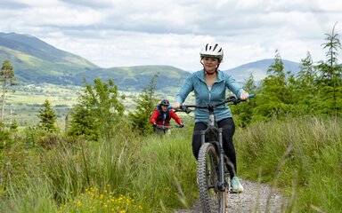 A woman in blue riding a mountain bike along a forest trail followed by a man in red