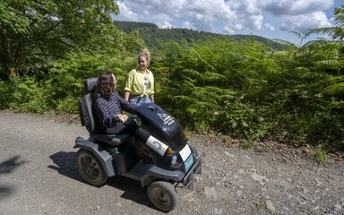 A youg woman on an off-road mobility scooter travels along a forest road whilst in conversation with another woman wearing yellow.