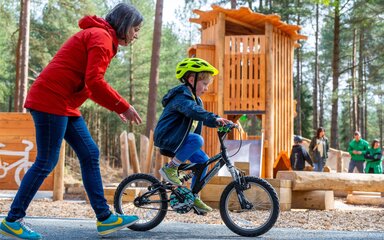 woman helping young child cycle with a wooden play area and forest background