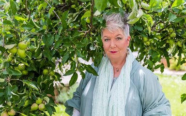A woman with short grey hair, fabulous shade of pink lipstick with long earrings stands by an apple tree. She wears a long pale green scarf over a long green dress.