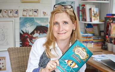 A woman with collar bone length blonde hair with glasses on her head holds a book she has written called How to train your dragon.