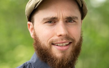 A colour photo of a man with a beard wearing a hat looking straight at the camera