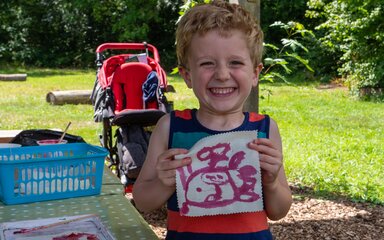 A young boy stands smiling at a piece of art work he has made on a piece of fabric.