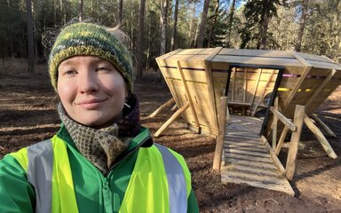 A woman in high-vis uniform taking a selfie by a wooden structure in the forest.