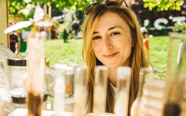 A lady with long blonde hair sis in front of a test tube rack. Sunglasses are placed on her head and she smiles at the camera.