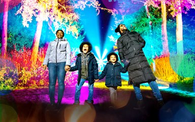 A family hold hands in a line looking in awe at brightly coloured lights of enchanted Chrismtas surrounding them.
