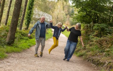 A family walking on a wide forest trail. The couple are swinging their daughter in the air.