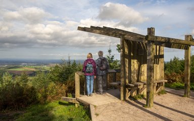 A couple stand under a wooden shelter looking out from a viewpoint across the countryside.