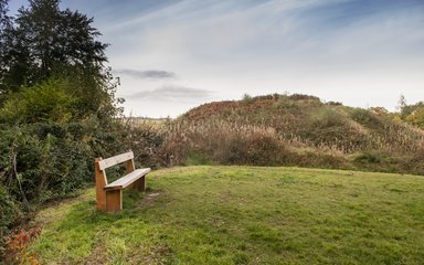 A motte and bailey monument viewed with a grassy area and wooden bench in the foreground