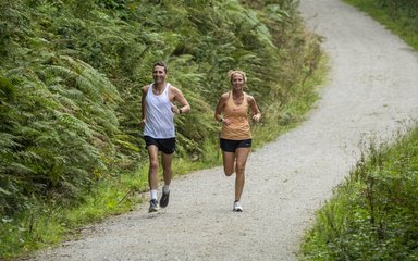 A male runner in a white vest and a female runner in an orange vest run towards the camera along a forest road