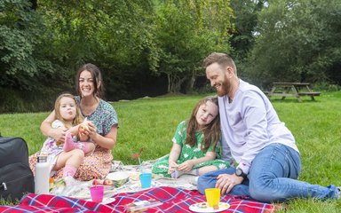 Two parents and two children sit on a tartan blanket having a picnic in a grassed area