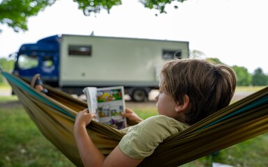 Boy reading a graphic novel whilst lying in a hammock, with a motorhome in the background